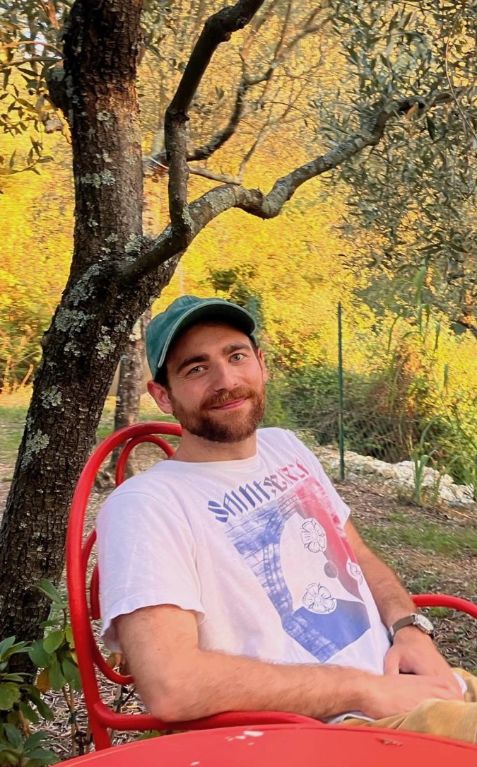 A picture of me, a man with brown hair and a short beard, wearing a baseball cap and a t-shirt, smiling, in northwestern Italy, in September 2023.
